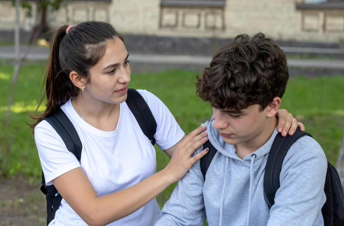 A person in a white t-shirt is gently consoling another person in a gray hoodie, who appears thoughtful and pensive. Both individuals are wearing backpacks, and they are outdoors in a grassy area with a building in the background.