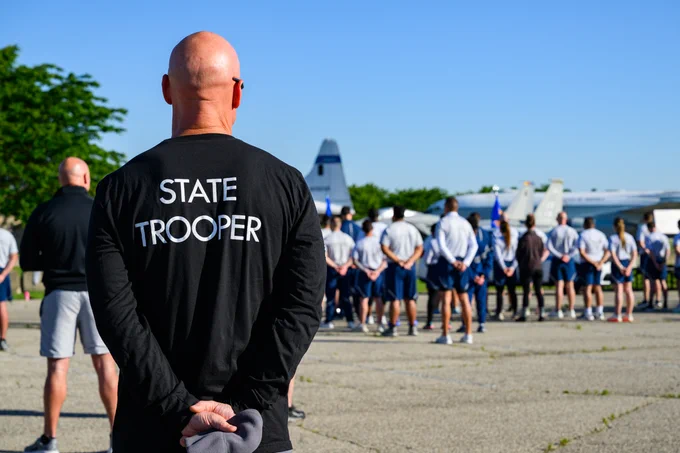 A person wearing a "State Trooper" shirt stands with hands behind their back, observing a group of people lined up in formation.