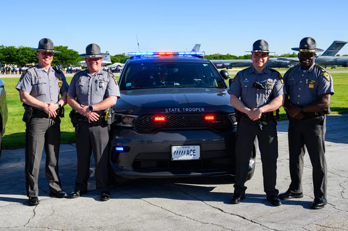 Four state troopers are standing in front of a state trooper cruiser with flashing lights. 