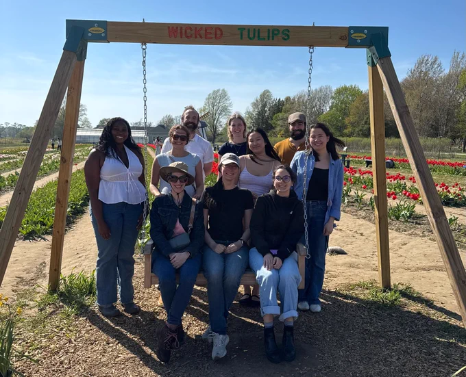 Lab members (left to right, front to back) Mmesoma Nwokolo, Theresa Desrochers, Hannah Hyde (née Doyle), Brent Stewart, Kati Conen, Ellie McVeigh, Aiai Calmer, Defne Buyukyazgan, Matthew Maestri, and Nami Kaneko all under a frame for a wooden swing labeled Wicked Tulips with Theresa, Kati, and Defne sitting in the swing. In the background are rows of planted tulips and trees at the edge of the field under a blue sky