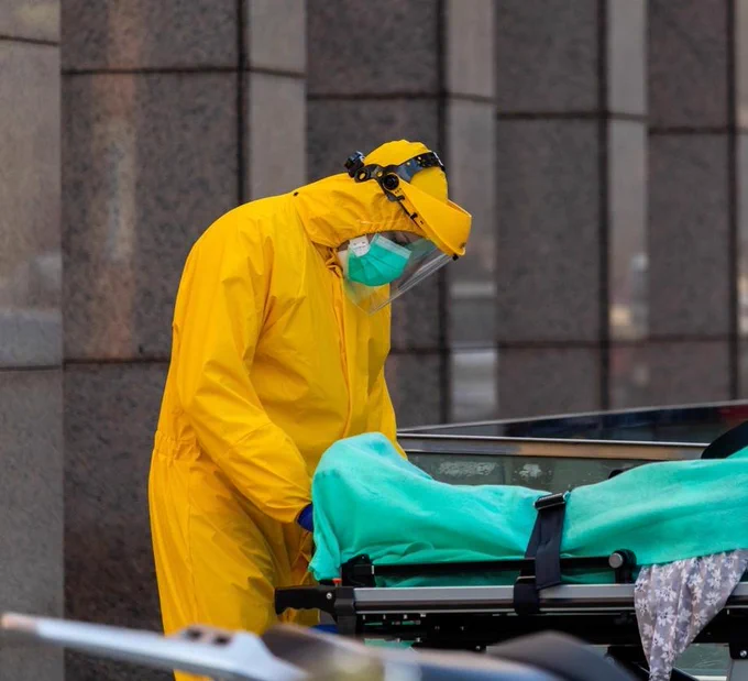 Healthcare worker in full yellow protective gear and face shield attending to a patient covered with a green blanket on a stretcher outside a building.