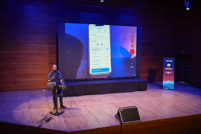 Wide shot of the Dani  presenting at iOSKonf in Skopje, with a large screen behind him displaying the camera feed a mobile app accessibility demo running on his device. The app is running VoiceOver. A conference banner is visible to the right with the name of talk (Accessibility, live on Stage) and the name of the speaker (Daniel Devesa Derksen-Staats).