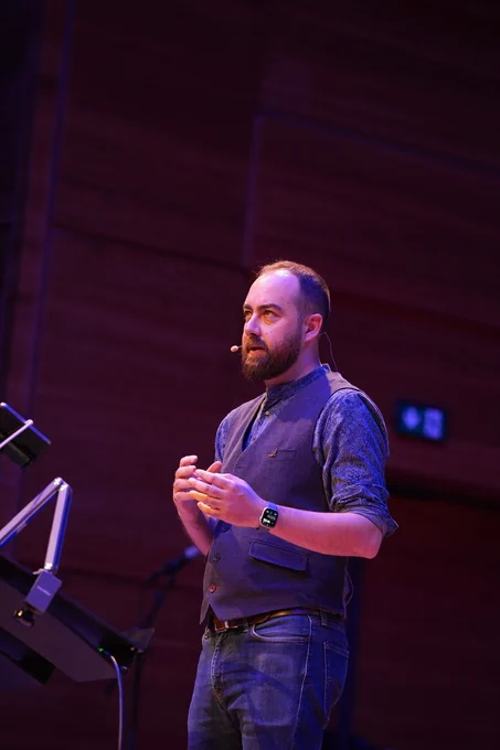 Close-up of Dani speaking at iOSKonf, wearing a dark blue vest and headset mic, hands clasped while speaking passionately on stage.
