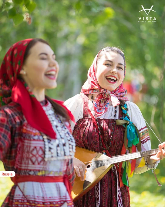 Two individuals in traditional Russian attire are joyfully singing and playing a balalaika in an outdoor setting. The background is filled with lush greenery, creating a vibrant atmosphere. The corner displays a small label reading "Russia," and the top right has the Vista logo.
