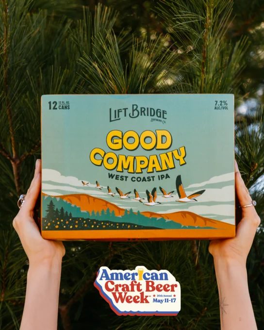 A person holds a box of Lift Bridge Brewing Co.'s Good Company West Coast IPA in front of a backdrop of lush green pine trees. The image promotes American Craft Beer Week, capturing a rustic, celebratory mood.