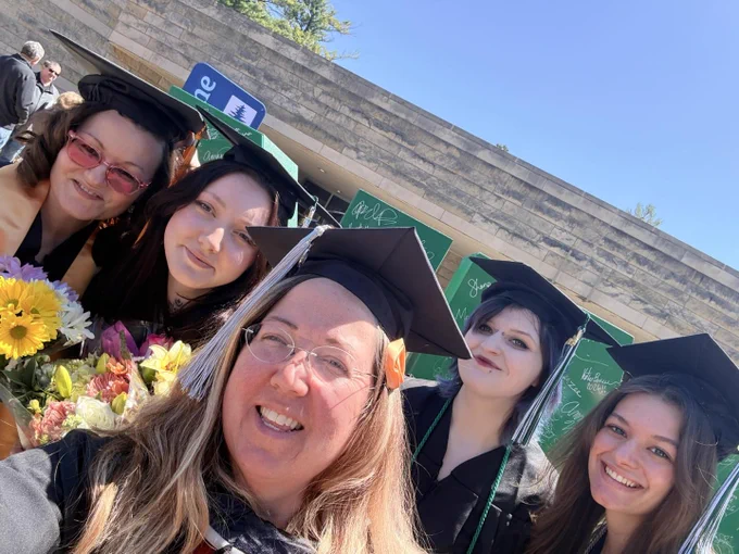 A group of five individuals wearing graduation caps and gowns are smiling. They are standing in front of a wall with green signs. One person is holding a bouquet of flowers. The scene is outdoors on a sunny day.