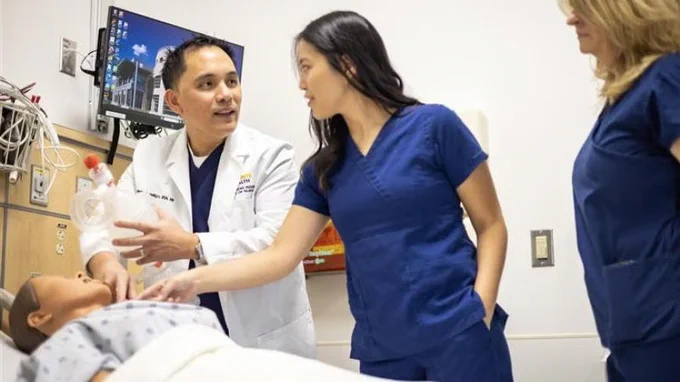 UC Davis Healthcare professionals from in a hospital room are working with a medical mannequin. One person in a white coat holds a medical tool, while two others in blue scrubs are watching and discussing. 