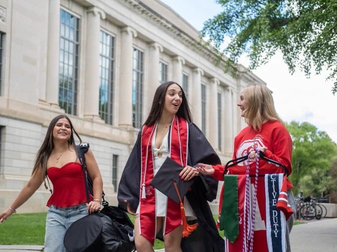 Three individuals in graduation attire walk together outside a large building on a college campus. They are smiling and holding academic regalia and flags with text including "ACTIVE DUTY." Trees and a clear sky are visible in the background.