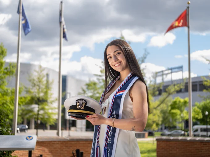 An individual outdoors holding a U.S. Navy officer hat, adorned with various sashes, and standing in front of a display with flagpoles in the background.