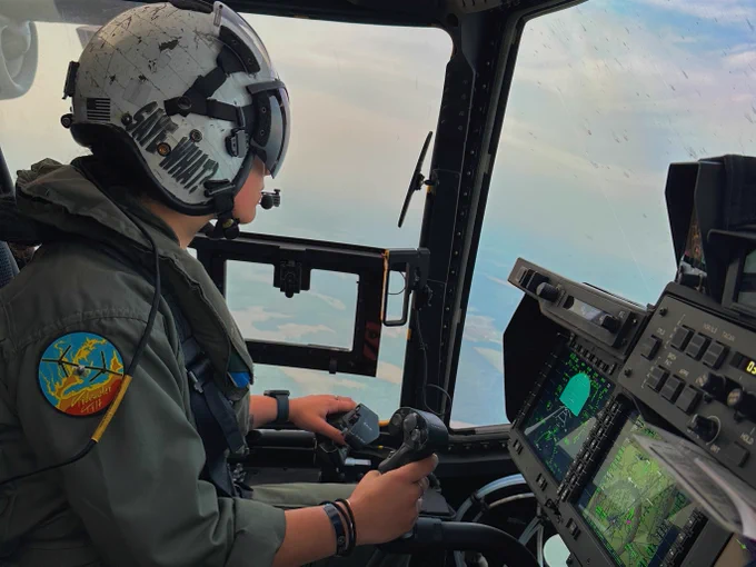 A person is seated in the cockpit of an aircraft wearing a helmet and flight suit. They are holding the controls while looking out the window. The cockpit is filled with various electronic displays and panels. A patch on the sleeve is visible.