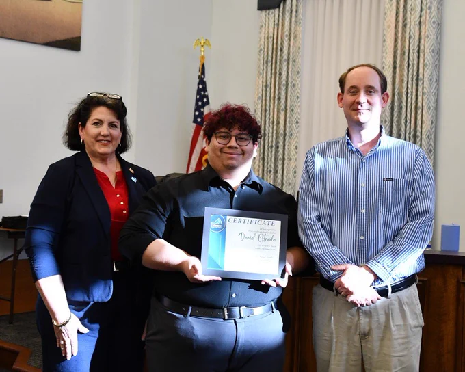 Three people stand in a room, one holding a framed certificate. Behind them, an American flag is visible.