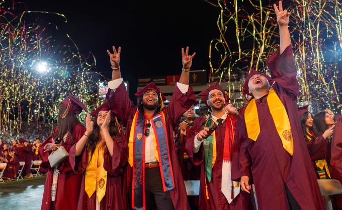 Group of graduates in maroon gowns celebrating with raised hands and confetti falling around them at night.