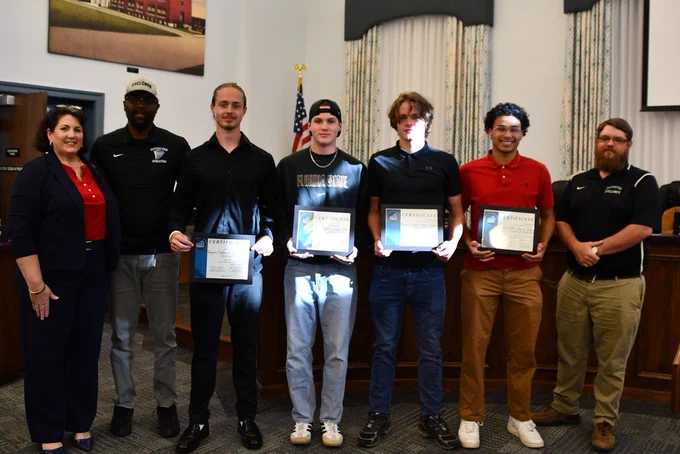 A group of seven people stands in a room, holding certificates, possibly at an award ceremony.