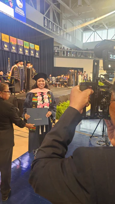 A female doctoral graduate poses for a photo with her degree cover. She is wearing a stole made of orchids and other pink flowers that match her doctoral hooding. She is also wearing a multicultural stole.