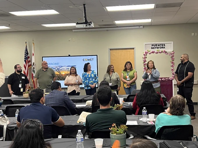 Facilitators and organizers stand at the front of a conference room during a Puentes Network COMET training while participants listen from their seats. A presentation screen and a floral “Puentes Network” frame are displayed behind them.