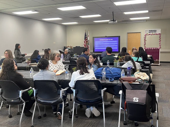 Participants sit around tables in a conference room during a Puentes Network COMET training session, speaking to each other.