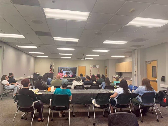 Participants sit around tables in a conference room during a Puentes Network COMET training session, listening to a presenter speaking in front of a screen.