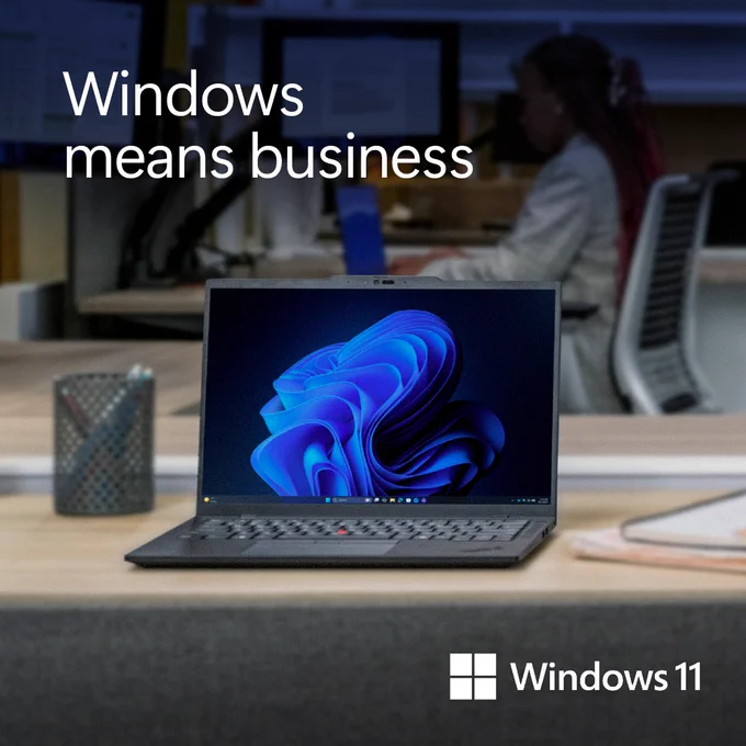 A Windows 11 laptop sits open on a desk in a modern office workspace, displaying the blue Windows 11 swirl wallpaper. In the background, an office workstation with a person seated at a computer is softly out of focus. On-screen text reads “Windows means business,” with the Windows 11 logo in the bottom corner.