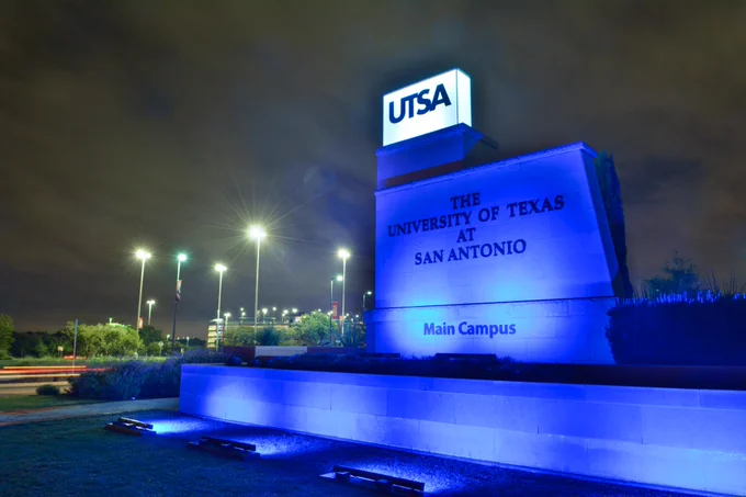 The image shows an illuminated sign for the University of Texas at San Antonio, Main Campus, glowing in blue light against a backdrop of night sky and streetlights.