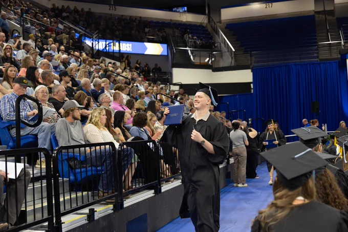 A graduate in a cap and gown holds a diploma while walking across a blue-floored arena stage, smiling. In the background, people fill the stadium seating, watching the ceremony.
