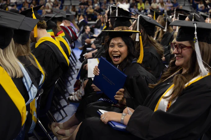 A seated group at a graduation ceremony, wearing black caps and gowns with yellow accents, holding diplomas. One person is smiling broadly at the camera, surrounded by others who are also celebrating. The setting appears to be an indoor auditorium.