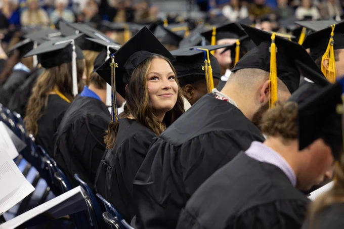 A group of graduates wearing black caps and gowns is seated in rows during a commencement ceremony. One person is looking upward with a smile, standing out from others who are focused on programs in their laps. The atmosphere is celebratory and formal.