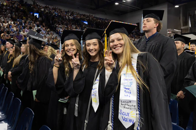 A group of people in graduation attire stand together inside a large venue. Three individuals at the front make a "SDState" Jackrabbit sign with their hands. One wears a decorated stole with the words "Sioux Valley" visible. The background is filled with more people and attendees.