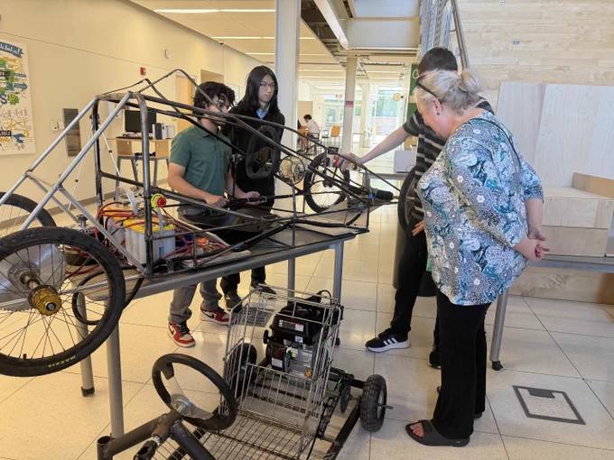 A person standing next to a CAPS Engineering cart on top of a table and a smaller one below it, while three students are explaining their projects.