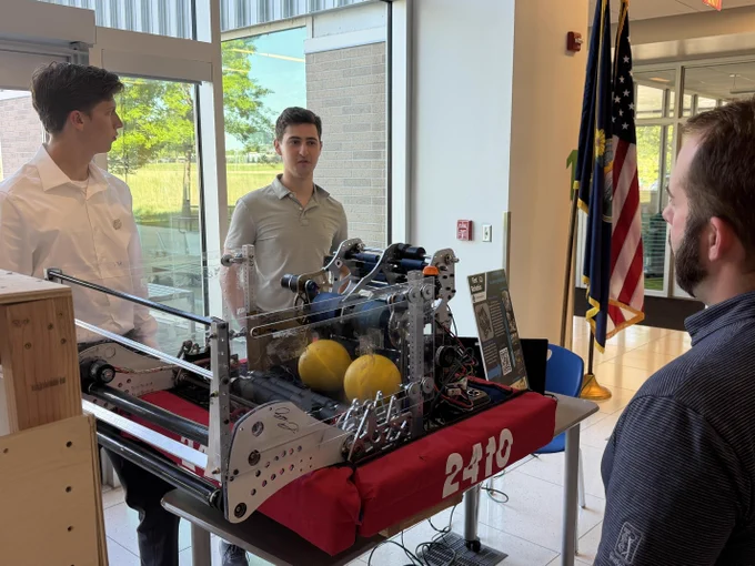 Several men standing behind a table displaying various tools and equipment, including a red robot. An American flag is visible in the background.