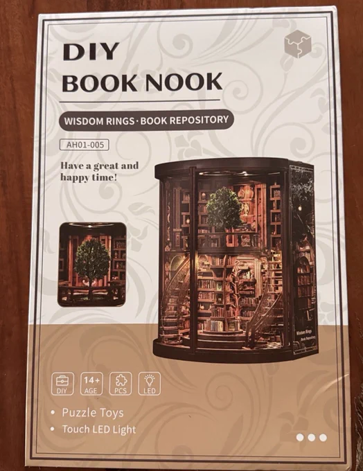 Verpackung eines DIY-Book-Nook-Bausets mit dem Titel „Wisdom Rings – Book Repository“. Abgebildet ist eine detailreiche Miniaturbibliothek mit geschwungenen Treppen, vielen Bücherregalen und einem großen Baum in der Mitte. Das Set enthält Puzzle-Teile und eine Touch-LED-Beleuchtung und ist für Personen ab 14 Jahren geeignet.