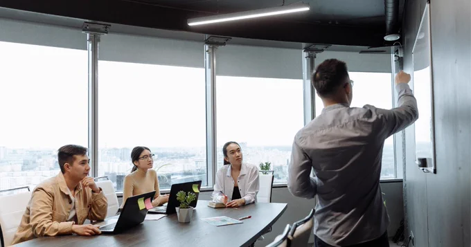 A person stands at a whiteboard presenting to three individuals seated at a conference table with laptops and notepads. The room has large windows with a view of a cityscape.