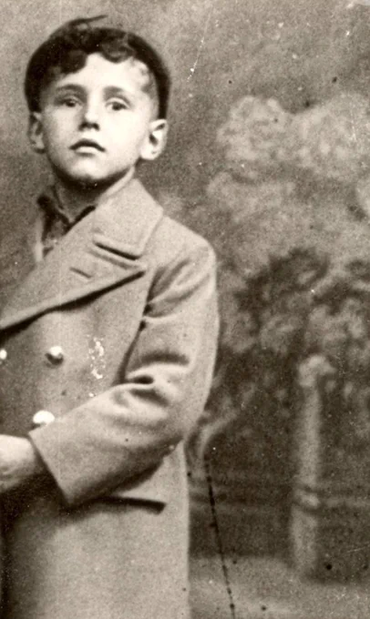 A young boy in a cap and coat photograph against a studio background with some fence and flowers.