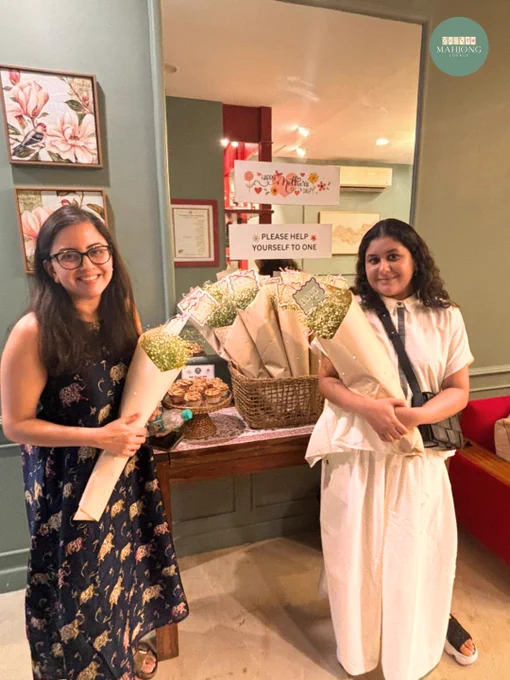 A cozy Mother’s Day celebration setup at Mahjong Lounge in Koramangala, Bangalore featuring women holding flower bouquets beside a beautifully styled Mahjong-themed table display. The warm editorial-style interiors include soft ambient lighting, elegant floral wall art, cupcakes arranged on woven stands, and baskets filled with wrapped bouquets decorated with Mahjong-themed tags. Signs reading “Happy Mother’s Day” and “Please Help Yourself To One” create a welcoming and thoughtful atmosphere. The women are smiling and standing inside a calm café-style Mahjong lounge space designed for community, connection, and meaningful offline experiences. The overall aesthetic blends warm tones, intimate hospitality, and luxury social club energy, highlighting Mahjong Lounge as a beginner-friendly Mahjong café and community hub in Bangalore where guests gather for Mahjong classes, social evenings, game nights, and shared moments with friends and family.