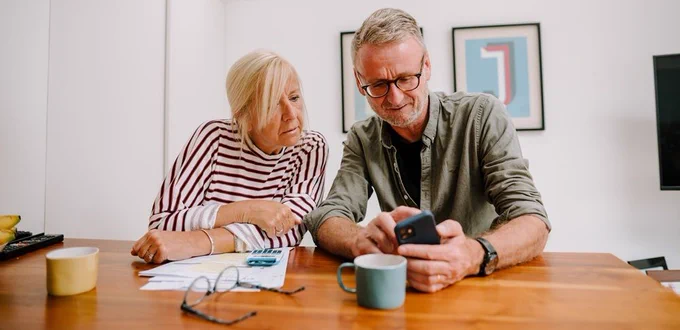 A male and female sitting side by side at a kitchen table. The male is looking at a phone whilst the female leans in to see what he is looking at.