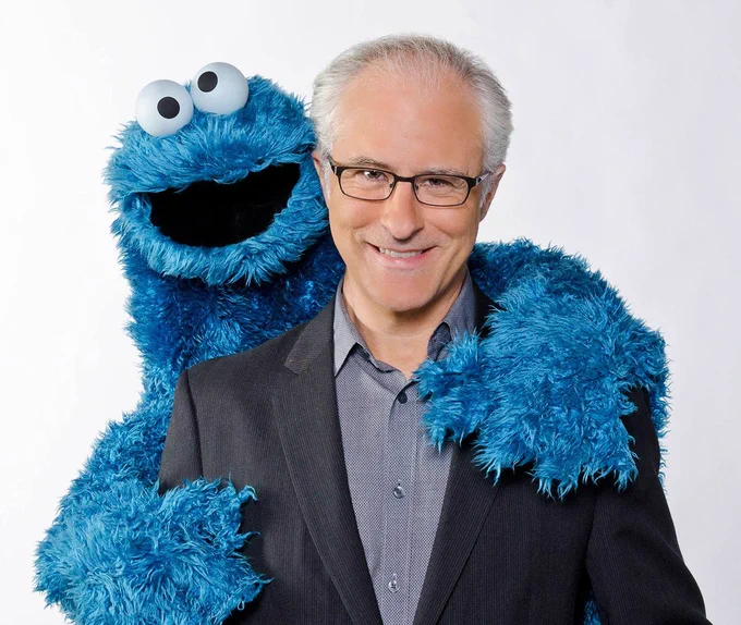 David Rudman, wearing a light blue button-down shirt and black jacket, smiles as Cookie Monster hugs him from behind. (Image courtesy of Muppet Wiki)