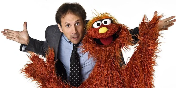 Joey Mazzarino, wearing a blue shirt, gray jacket, and black patterned tie shrugs as he stands behind Murray Monster. (Image courtesy of Muppet Wiki)