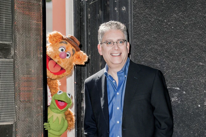 Bill Prady, wearing a blue button-down shirt and dark blue jacket, smiles near as Fozzie Bear and Kermit peer in from behind a studio door. (Image courtesy of Muppet Wiki)