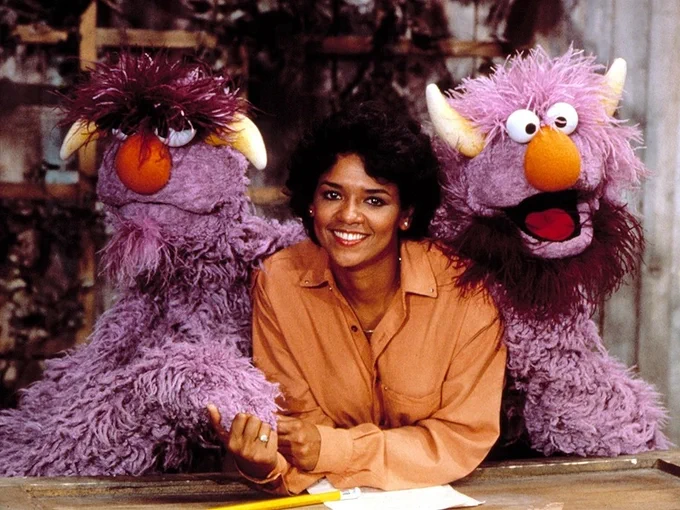 Sonia Manzano, as Maria, smiles in the courtyard wearing an orange shirt, as the Two-Headed Monster surrounds her. (Image courtesy of Muppet Wiki)