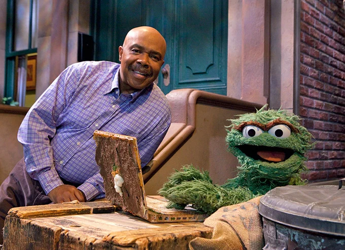 Roscoe Orman, as Gordon, stands on the front steps of 123 Sesame Street, smiling as he looks over Oscar the Grouch using a muddy laptop. (Image courtesy of Muppet Wiki)