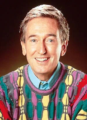 Bob McGrath, wearing a black, purple, red, green, and yellow sweater with a light blue shirt, smiles on a dark orange background. (Image courtesy of Muppet Wiki)
