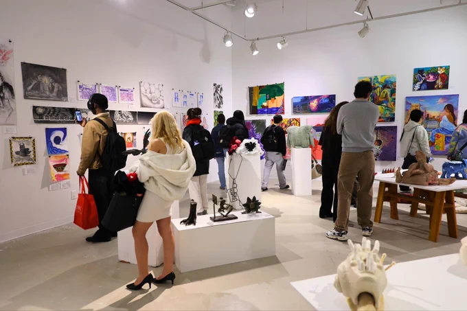 A busy art gallery with visitors observing various paintings and sculptures. The walls display colorful artworks, and several sculptures are placed on white pedestals. Some visitors are taking photos, while others engage with the art pieces. The gallery is well-lit with ceiling spotlights.