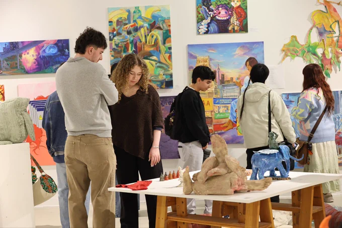 A group of people observe various colorful artworks in a gallery. The room features abstract paintings and sculptures, with several pieces displayed on walls and tables.