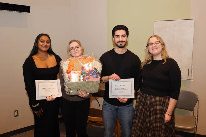 Four individuals standing indoors, smiling, and holding certificates. One person in the center is holding a large gift basket.