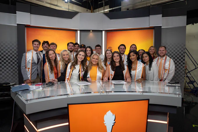 A group of smiling graduates wearing stoles poses behind a sleek news desk in a modern studio. The vibrant backdrop features orange tones, adding warmth and energy.