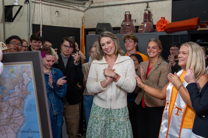 A joyful group of people, including a woman in a white cardigan and green skirt, smiling and clapping in a room with industrial equipment and a map.