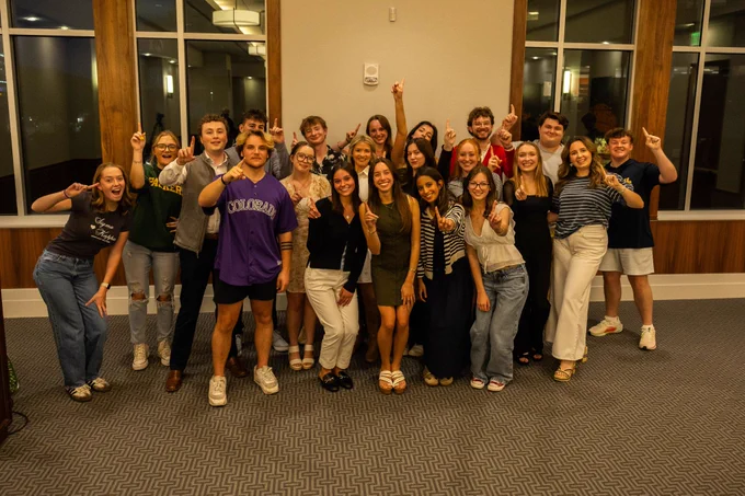 A diverse group of 24 smiling young adults, some pointing upwards, gather indoors against a windowed wall, conveying a joyful, celebratory tone.