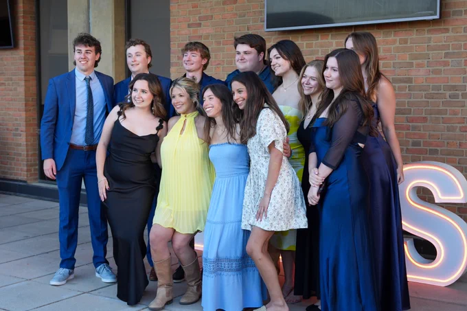 A group of young people, dressed formally in suits and colorful dresses, pose cheerfully together in front of a brick wall and illuminated letters.