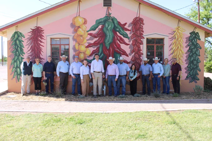 A group of people stands in front of a brightly painted building featuring large murals of chili peppers. The individuals are smiling, wearing a mix of casual and formal attire, including cowboy hats. The scene takes place on a sunny day, with clear skies visible behind the building.