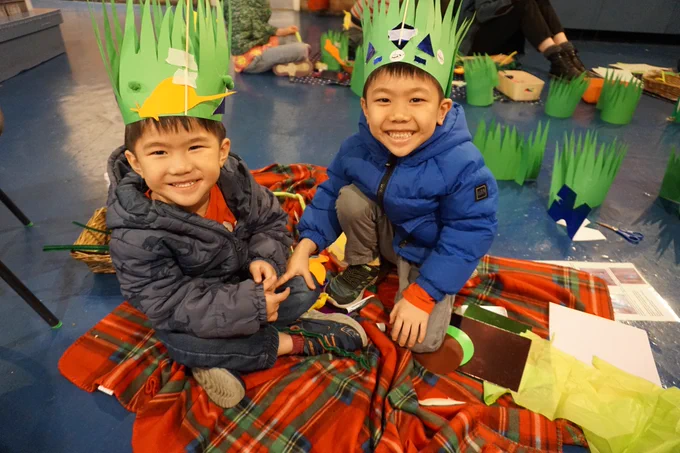 Two young children sit together on a tartan blanket inside the London Canal Museum, both wearing handmade green nature crowns decorated with paper wildlife shapes, smiling at the camera.
