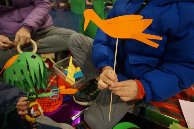 A child holds up a paper bird — cut from orange card and mounted on a wooden stick — during a craft activity at the London Canal Museum. Nearby, another child holds a green paper crown decorated with orange and purple shapes. A tray of coloured paper cut-outs, scissors, and pipe cleaners sits between them on the floor.